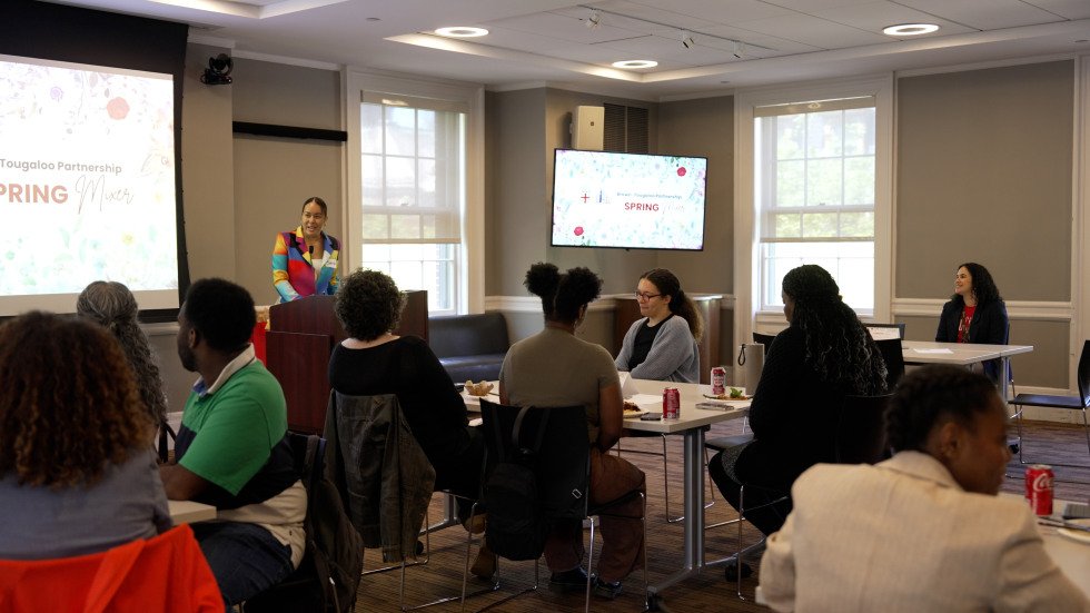 A group of students, faculty, and staff watch a female presenter share remarks in front of a slide. 