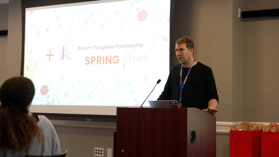 A male presenter shares remarks in front of a slide. 