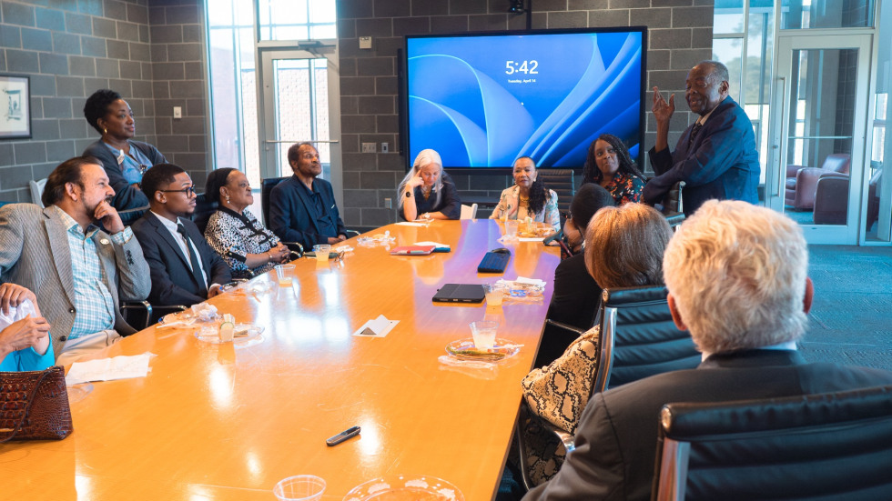 A group of people sit around a conference room table listening to a man who is standing up sharing remarks