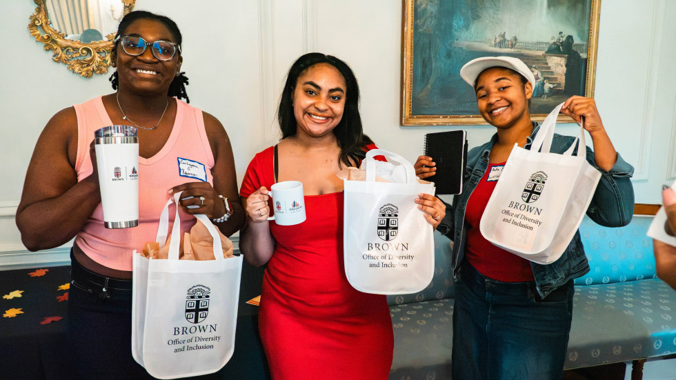 Three students hold white bags that have the Brown Crest and "Office of Diversity and Inclusion", and mugs that say Brown-Tougaloo Partnership